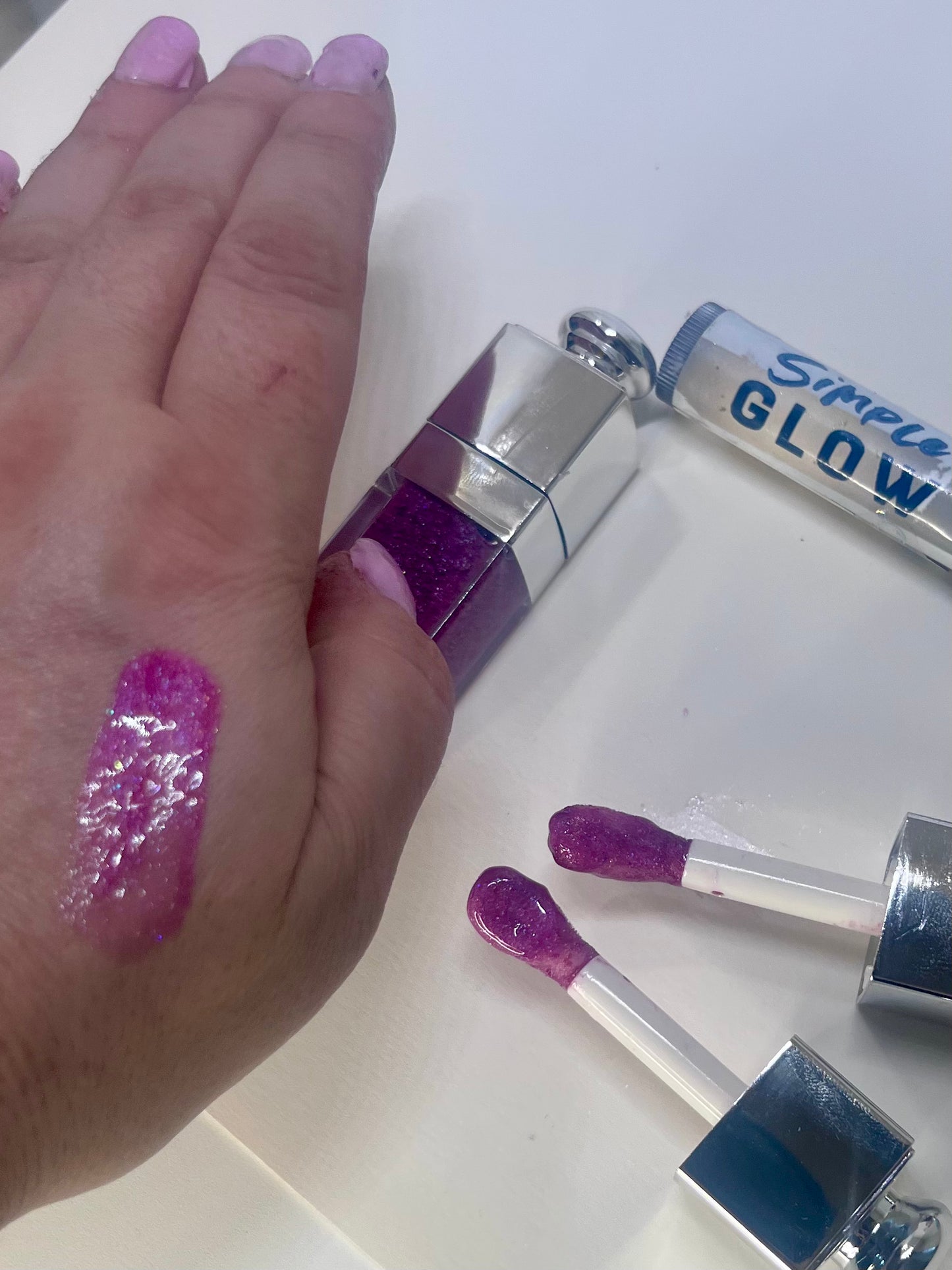 Hand with mauve glittery lip gloss swatch and lip gloss applicators on a white background