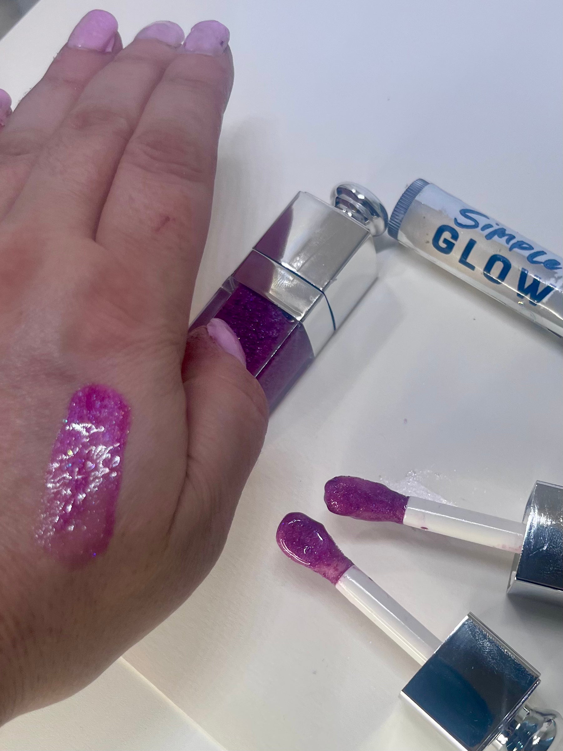 Hand with mauve glittery lip gloss swatch and lip gloss applicators on a white background