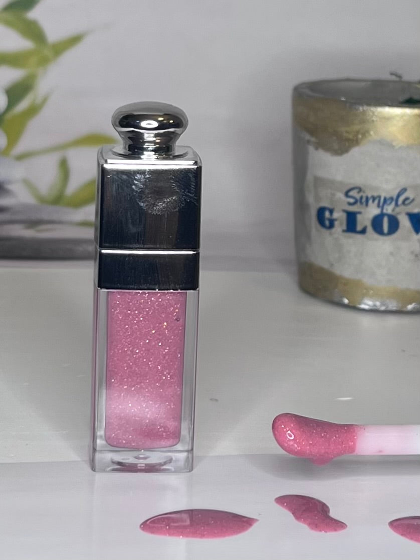Pink glitter lip gloss with silver cap on a white surface, with 'Simple Glow' product in the background.